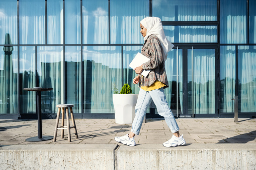 mujer joven con hiyab caminando con computadora portátil frente a la fachada de vidrio photo