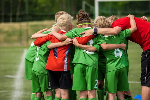 Photo of Happy sports team with a young coach. Children playing a football game on stadium pitch. Trainer motivating young players during the pregame briefing. Boys in green soccer jersey uniforms