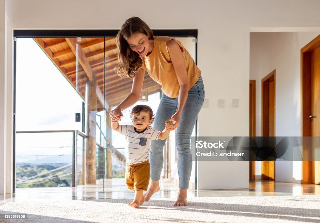 Toddler learning how to walk at home with the help of his mother Happy toddler at home learning how to walk with the help of his mother - first steps concepts Family Stock Photo