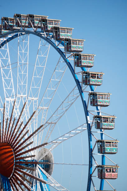 A large Ferris wheel with empty cabins against a cloudy sky, partially obscured by a wooden building.