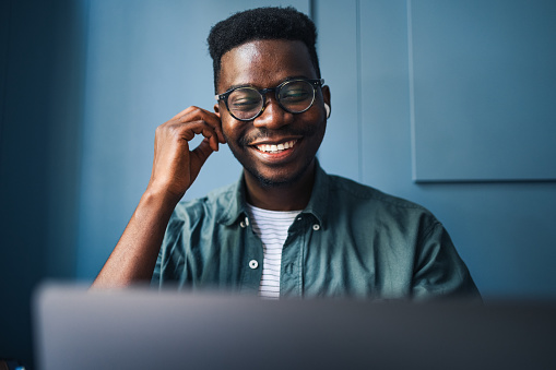 Smiling African-American man with wireless earphones listening online lessons using a laptop computer from a cafe.