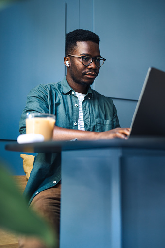 Serious African-American man with wireless earphones listening online lessons and typing notes on a laptop keyboard from a cafe.