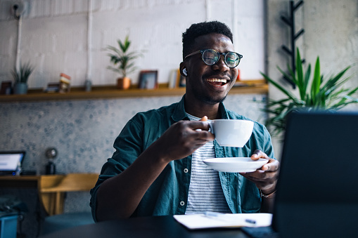 Cheerful smiling African-American man with bluetooth earphones listening online lessons on his laptop while drinking cup of coffee in a cafe.