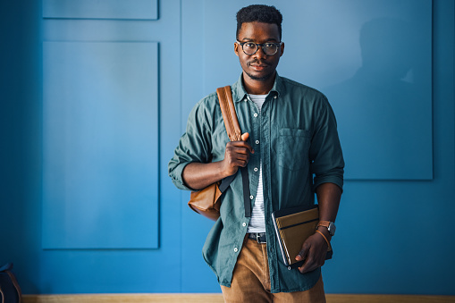 Serious African-American student with eyeglasses looking in camera and carrying a backpack and notebooks in a cafe.