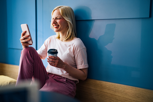 Cheerful smiling young woman using her smartphone while sitting in a coffee shop with disposable cup of coffee in hands.