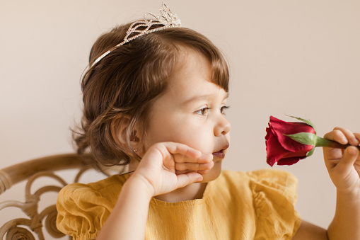 An Almost 3-Year-Old Toddler Girl Wearing a Yellow-Colored Dress & a Tiara Looking At a Red Rose.