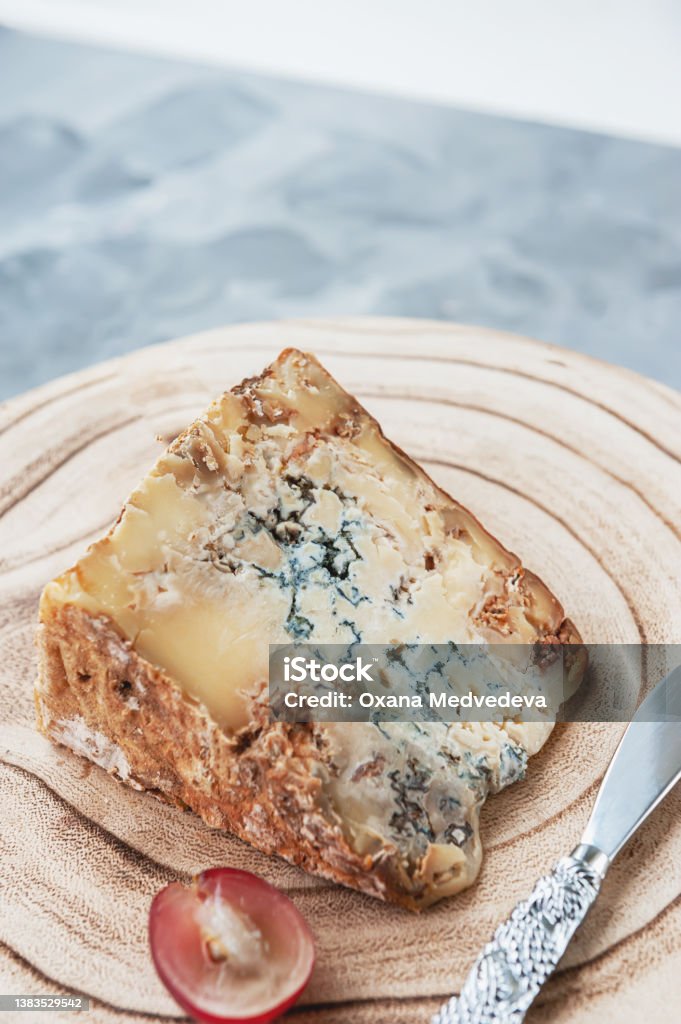 large piece of stilton cheese is lying on a wooden board. Delicacy cheese with blue mold stilton with grapes. large piece of stilton cheese is lying on a wooden board. Delicacy cheese with blue mold stilton with grapes. Copy space. Stilton Cheese Stock Photo