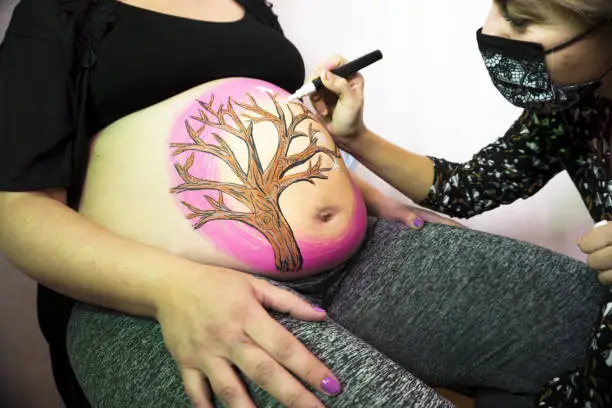 Body painting session on the belly of an eight month pregnant woman. Drawing of a tree