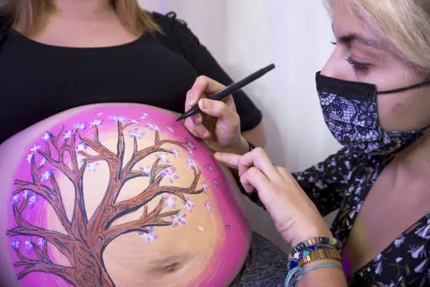 Body painting session on the belly of an eight month pregnant woman. Drawing of a tree