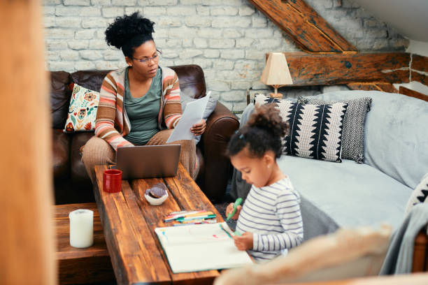 istock Black working mother analyzing paperwork and using laptop while her daughter is coloring on the paper at home. 1383532083