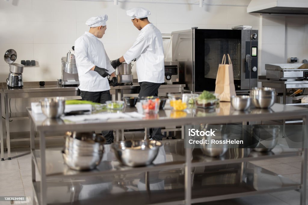 Two chef cooks standing together in professional kitchen Two chef cooks standing together in professional kitchen with food ingredients in front. Latin and Asian guy working together as cooks at restaurant Adult Stock Photo