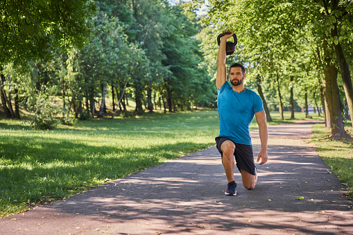 Young man exercises kettlebell lunges during a workout outside in a park during sunny summer day