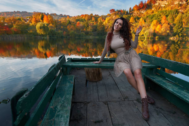Beautiful fashion model sitting on a jetty leading into a lake surrounded by colorful forests reflecting in the water