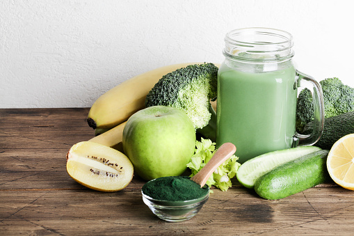 green smoothie in glass bottle, spirulina powder, vegetables and fruits on wooden background. healthy, raw, vegan diet concept. copy space