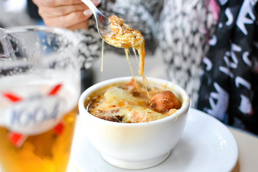 A woman sits at an outdoor sidewalk cafe in Paris France eating a bowl of French Onion Soup on a spoon with a 1664 beer.