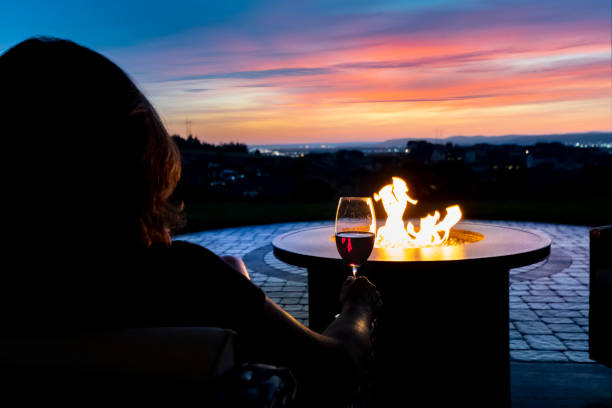 una donna si rilassa con un bicchiere di vino rosso da un pozzo del fuoco nel patio di una casa di lusso che si affaccia su una valle e luci della città dello skyline durante un colorato tramonto rosa, giallo e blu. - fire pit foto e immagini stock