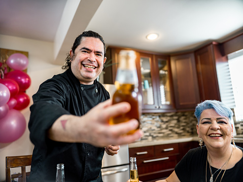 LGBTQ Mexican Male Chef serving beer and disabled mature woman at the party table with friends and family. He is dressed in black chefs robe, she is dressed in casual clothes. Interior of kitchen in private city home.