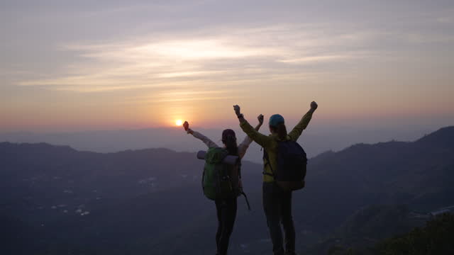 Two woman hiking to the mountain and giving high fives on top of mountain and raising her hands celebrating successful