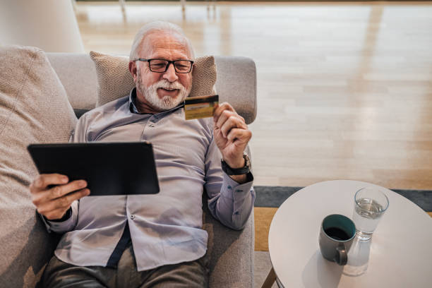 istock Senior good-looking handsome man lying down on sofa or couch at his home or office using hand held tablet device and credit card for online shopping web paying, buying presents for friends and family 1383570336