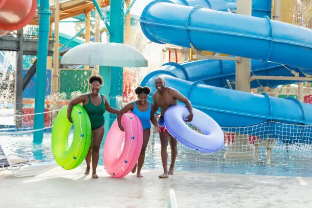 An African-American family with a 12 year old girl having fun at a water park. They are carrying inflatable rings, ready to float on the lazy river, laughing and looking toward the camera. Giant water slides are in the background.