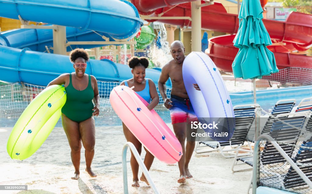 Family at waterpark carry inflatable rings to lazy river An African-American family with a 12 year old girl having fun at a water park. They are carrying inflatable rings, ready to float on the lazy river. Giant water slides are in the background. Water Park Stock Photo