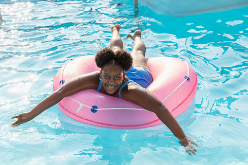 A 12 year old African-American girl having fun at a water park, floating on a lazy river on a pink inflatable ring. She is lying on her front, smiling at the camera.