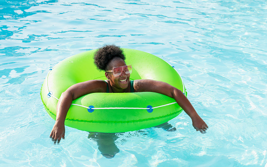 A mature African-American woman in her 40s having fun at a water park, floating on an inflatable ring on the lazy river. She is smiling, looking at the camera with a sideways glance.