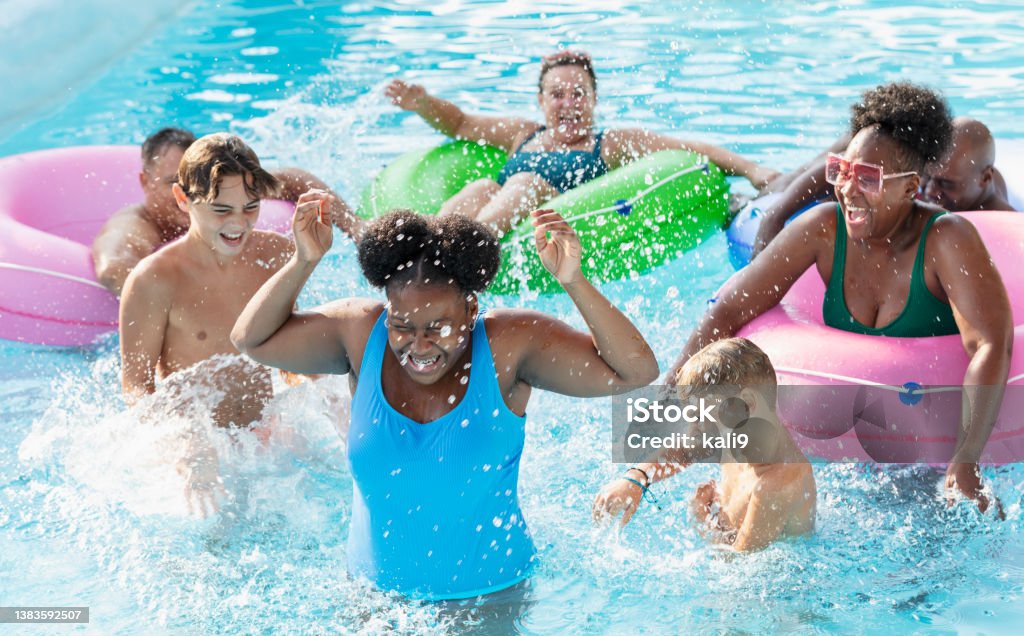 Multiracial friends and family splashing on lazy river A multiracial group of seven people, two families, having fun at a water park on the lazy river. The four parents are floating in inflatable rings. The three children are standing waist deep in the water. The 12 year old African-American girl in the foreground is laughing with her eyes closed as she gets splashed. Swimming Pool Stock Photo