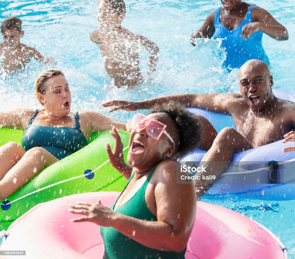 Multiracial friends and family splashing on lazy river A multiracial group of six people, two families, having fun at a water park on the lazy river. The three parents are floating in inflatable rings. The three children are standing waist deep in the water behind them. The African-American woman in the foreground is laughing as she gets splashed. Family Stock Photo