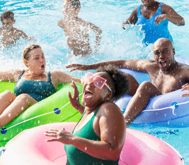 Multiracial friends and family splashing on lazy river A multiracial group of six people, two families, having fun at a water park on the lazy river. The three parents are floating in inflatable rings. The three children are standing waist deep in the water behind them. The African-American woman in the foreground is laughing as she gets splashed. inflatable ring photos stock pictures, royalty-free photos & images