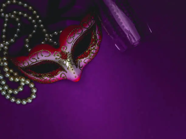 A masquerade mask, white pearl beads and an empty champagne glass lie on the left on a purple-lilac background with copy space on the right, flat lay close-up.Mardi gras celebration concept.