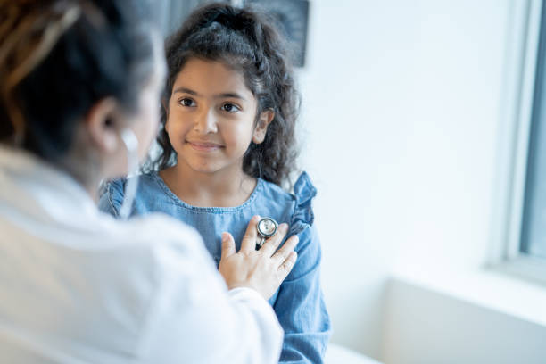 Doctor Listening to a Young Girls Heart A female doctor of Latin decent, holds her stethoscope to her young patients chest as she listens to her heartbeat while she sits up on an exam table. The doctor is wearing a white lab coat and the patient is dressed casually as she looks up at her doctor. general practitioner stock pictures, royalty-free photos & images