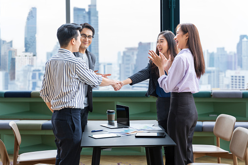 Asian business team leader congratulate his teammate employee for the outstanding achievement team performance by shaking hand in modern office workplace with skyscraper view