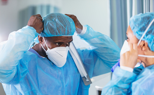 Two multiracial healthcare workers working in a hospital ward, putting on full PPE. They are wearing surgical caps and gowns and adjusting their N95 face masks. The woman is mixed race African-American and White. The focus is on her coworker, an African-American man. Both are in their 40s. This is part of a series showing healthcare workers in various stages of putting on or working in PPE (personal protective equipment).