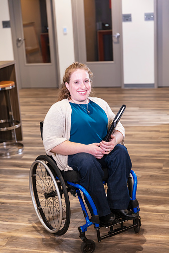 A mid adult woman working in an office, using a self-propelled wheelchair. She is holding a file folder, smiling confidently at the camera.