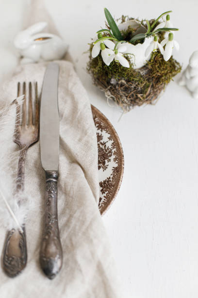 istock Stylish Easter table setting. Natural egg with snowdrops in nest, feathers on napkin on modern plate, cutlery, bunny and  on white wooden table. Modern Easter table rustic decoration 1383792085