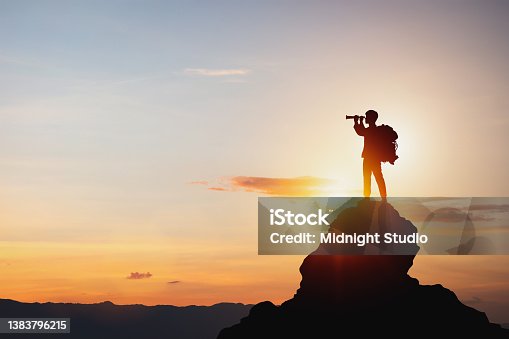 istock Silhouette of man holding binoculars on mountain peak against bright sunlight sky background. 1383796215