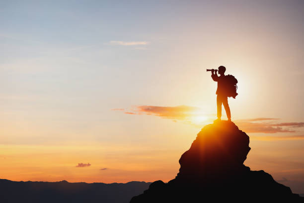 Photo of Silhouette of man holding binoculars on mountain peak against bright sunlight sky background.
