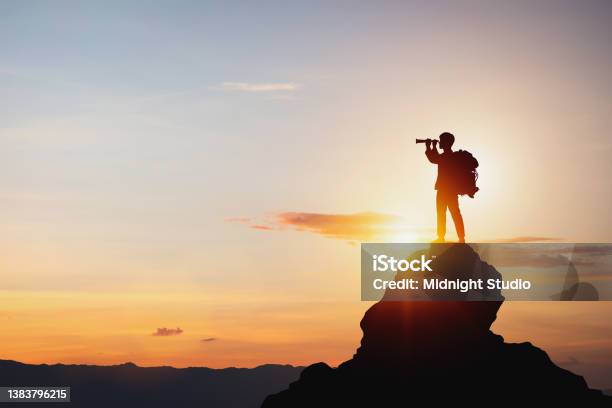 Silhouette Of Man Holding Binoculars On Mountain Peak Against Bright Sunlight Sky Background Stock Photo - Download Image Now