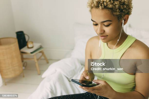 Close up of dark skinned dyed white hair teenage girl with natural beauty wearing body jewelry sitting on bed in earphones choosing music in smartphone for gymnastics at home. Domestic sports concept