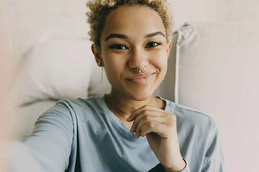 Close-up of cute African ethnicity girl recording stories for her blog using selfie camera of her smartphone, sharing information, dressed in grey blue shirt posing against cosy home background