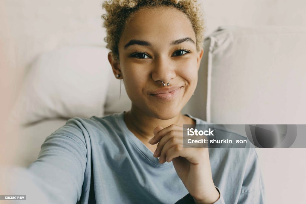 Close-up of cute African ethnicity girl recording stories for her blog using selfie camera of her smartphone, sharing information, dressed in grey blue shirt posing against cosy home background Nose Ring Stock Photo