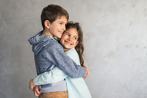 Smiling loving young boy and girl hugging. Portrait of them against grey wall. Copy space.