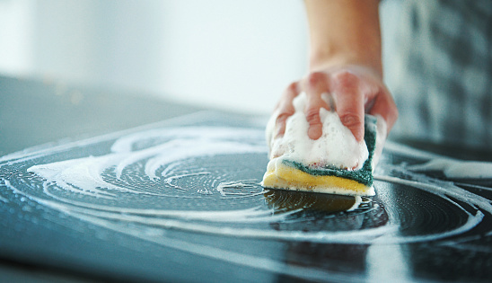 Closeup of unrecognizable woman cleaning glass ceramic stove top with a detergent and a soft cleaning sponge.
