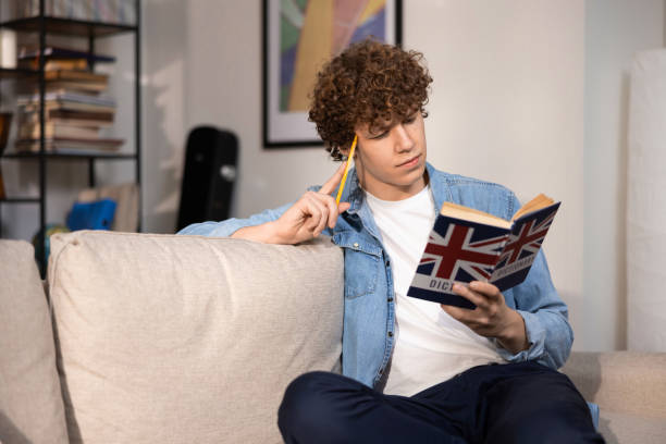 istock A handsome college student reads an English dictionary while sitting on the couch in the living room of his family's home. A teenage boy in a denim shirt repeats more new words in his mind 1383835233