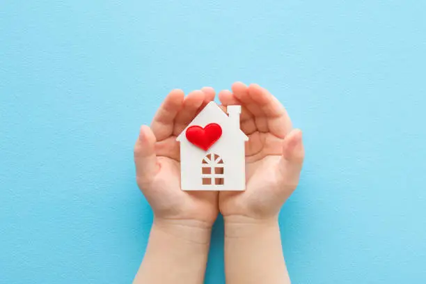 Baby opened palms holding white house with bright red heart on light blue table background. Pastel color. Closeup. Point of view shot. Concept of love and happiness at family home. Top down view.