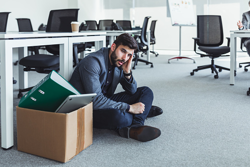 Dismissed millenial sitting on the office floor. Resignation letter in cardboard box and employee headache in office. Quitting a job, businessman fired or leave a job concept.