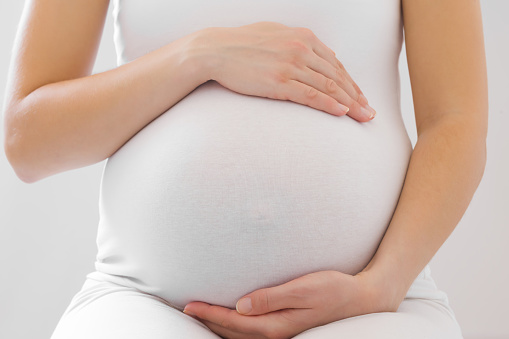 Young adult pregnant woman in white clothes touching big belly and feeling her baby move on light gray background. Pregnancy time concept. Expectation time. Closeup. Front view.