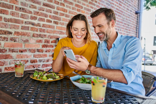Happy couple sharing smartphone while out in the city eating lunch at outdoors cafe stock photo