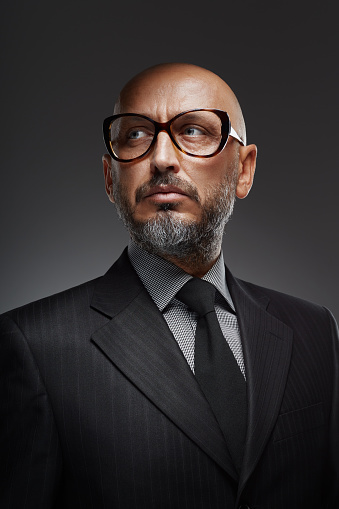 Studio portrait of a middle-aged confident and successful businessman with retro eyeglasses posing against a dark background